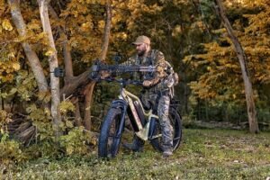 Metsästäjä maastokuvioisissa vaatteissa sähkö-fatbiken kanssa syksyisessä metsässä.