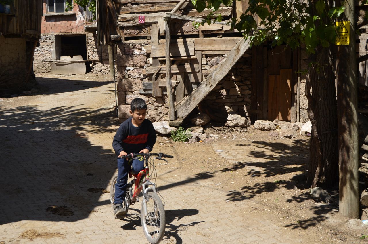 Young boy rides a bicycle through a rustic village street on a sunny day, enjoying outdoor leisure.