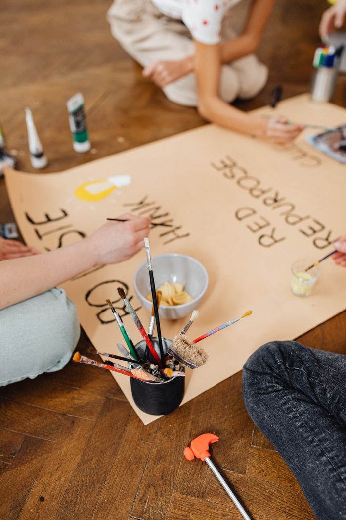 A close-up of hands painting a large paper on a wooden floor with brushes and paint.